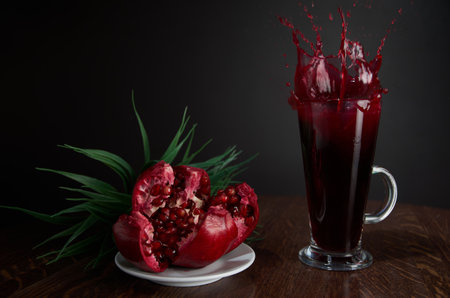 Red pomegranate juice splash in a beautiful glass, next to it lies a broken pomegranate on a wooden table with a dark background in the interior of the cafe. Drops juice scatter from the glass.の写真素材