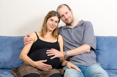 Smiling man embracing happy pregnant woman sitting on couch holding tummyのeditorial素材