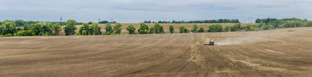 Panorama plowed field with tractors in agricultural work on the background of treesの写真素材