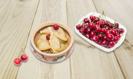 Ukrainian varenyky (dumplings) filled with sour cherry in ceramic bowl, transparent plastic tray with dark red cherries and two cherries separately on a wooden surfaceの写真素材