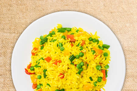 Fragment of white dish with boiled rice, cooked with turmeric, with boiled vegetables - carrots, bell peppers, green peas, green beans closeup on the surface of scrim.の写真素材