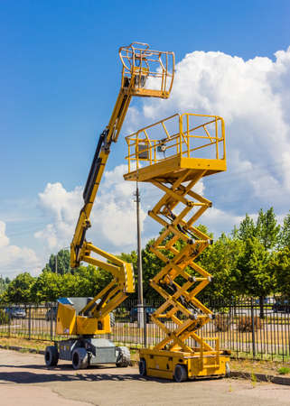 Two types of mobile aerial work platform - yellow scissor hydraulic lift and yellow hydraulic articulated boom lift against the sky with clouds and trees