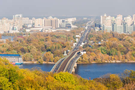 Autumn cityscape with yellowed leaves on the trees, the river and the bridge across it in the foreground, urban multi storey buildings and sky in the background. Kiev, Ukraine.の写真素材