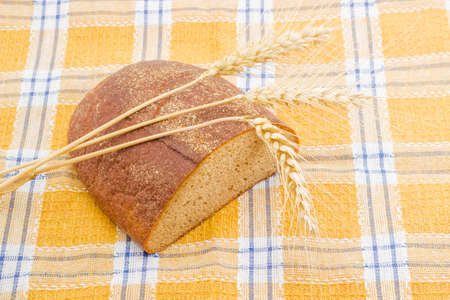 Half a oblong loaf of brown bread and three wheat spikes closeup on a checkered tablecloth.