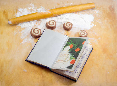 Rolling pin, flour, uncooked sweet roll, made from two color shortbread dough and notebook to write recipes on wooden cutting board during making sweet rollの写真素材