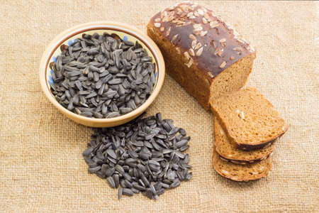 Partly sliced brown bread baked with kernels of a sunflower seeds, pile of a sunflower seeds and bowl with seeds not peeled  from the husk on the sackclothの写真素材