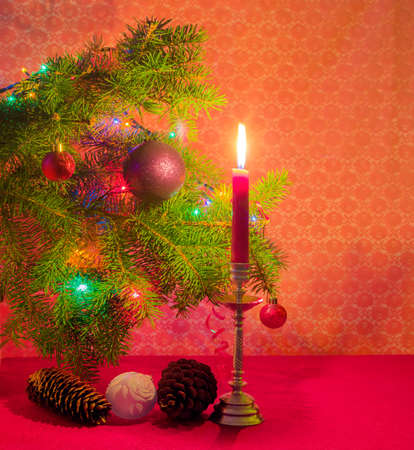 Branch of a Christmas trees, decorated with Christmas ornament and lights, fir and pine cones and candle in a candlestick in the light of candle on a red backgroundの写真素材