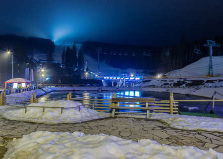 Night view to the wooded hillside with a ski pistes and chairlifts and a pond in the foreground at ski resortの写真素材