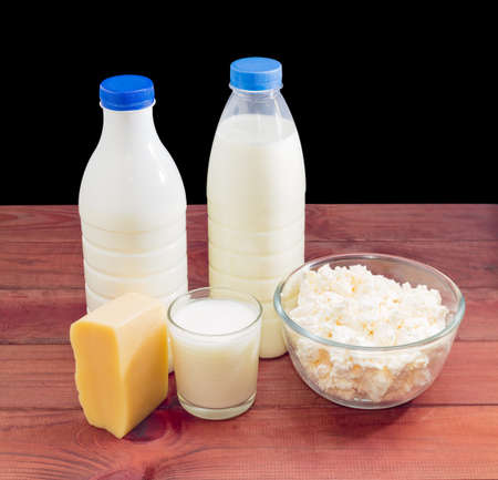 One plastic bottle with milk, one plastic bottle with fermented milk drink, glass bowl with cottage cheese, glass with milk and piece of a cheese on a dark wooden surfaceの写真素材