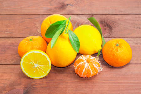 Several fresh whole mandarins, oranges and lemons with twigs and leaves, one lemon cut in half, one peeled mandarin on a dark wooden surfaceの写真素材