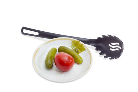 Two pickled cucumbers and one pickled tomato on a saucer and black plastic slotted spoon on a light backgroundの写真素材