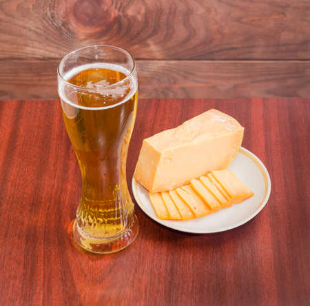 Beer glass with lager beer, one piece and several thin slices of hard cheese on saucer on wooden table against the backdrop of dark wooden planksの写真素材