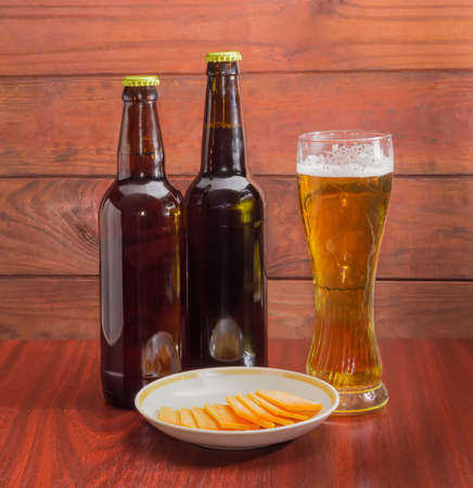 Glass of lager beer, two bottles of beer and several slices of cheese on saucer on wooden table against the backdrop of dark wooden planksの写真素材
