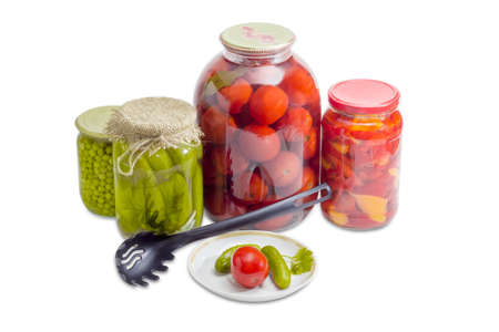 Two pickled cucumbers and one pickled tomato on a saucer, pickled cucumbers, tomatoes, bell pepper and green peas in glass jars and black plastic slotted spoon on a light backgroundの写真素材