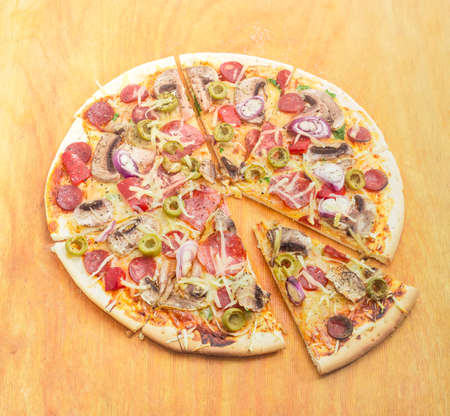 Sliced cooked round pizza with various sausages, button mushrooms and olives on wooden cutting board on a light backgroundの写真素材