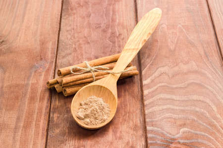 Several cinnamon sticks tied by twine and cinnamon powder in a wooden spoon on a surface with wooden planks closeupの写真素材