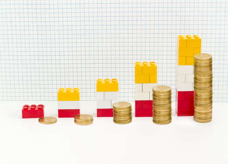 Stacked bar chart with bars which are made from colored plastic parts children's designer and stacks of coins against background of squared paperの写真素材