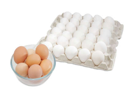 Fresh brown free-range eggs in a glass bowl and white chicken eggs in an egg tray from commercial factory farming on a light backgroundの写真素材