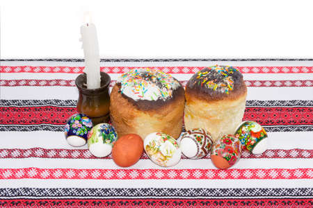 Two Easter cake decorated with white icing and colorful sugar decors, several Easter eggs with a various decorating and burning candle on a tablecloth with ornament on a light backgroundの写真素材