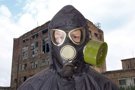 Head and shoulders of a person in a rubber gas mask and in a black jacket with a hood against the backdrop of abandoned and partially destroyed industrial buildingの写真素材