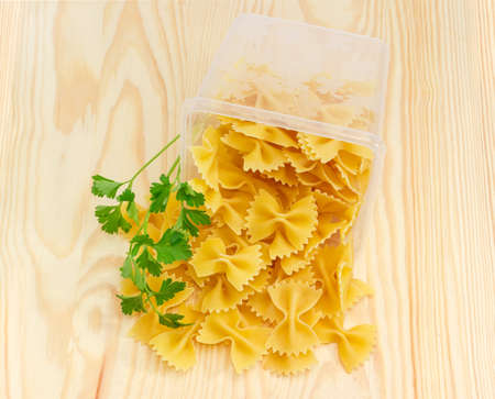 Uncooked bow-tie pasta in plastic tray and next to it and sprig of a parsley on a light wooden surfaceの写真素材