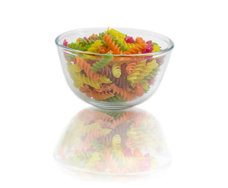 Uncooked dried spiral pasta colored by beetroot, spinach, paprika, carrot, grape seeds in a transparent glass bowlの写真素材