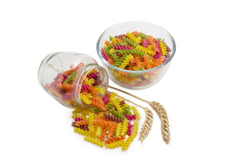 Uncooked dried spiral pasta colored by beetroot, spinach, paprika, carrot, grape seeds in a transparent glass bowl and jar, two wheat spikes on a light backgroundの写真素材