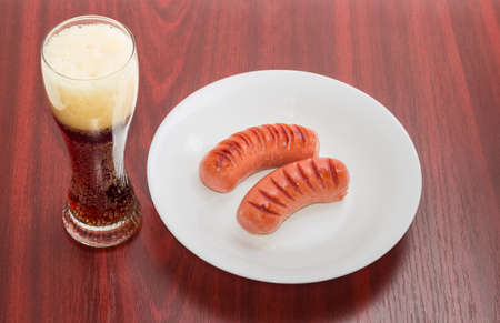 Two fried thick frankfurters on a white dish and glass of a dark beer on a wooden tableの写真素材