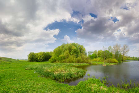 Spring landscape with lake, meadow on the coast and forest on the background of a sky with clouds in the afternoonの写真素材