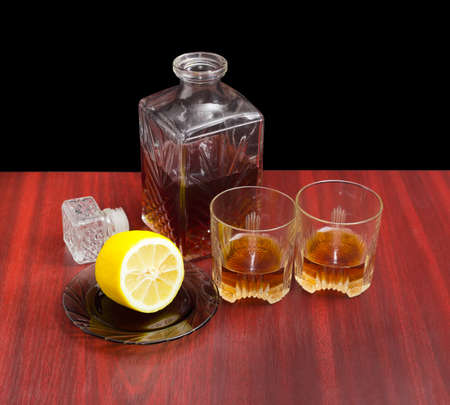 Decanter and two glasses with whiskey, stopper from the decanter and lemon on a saucer on a wooden table on a dark backgroundの写真素材