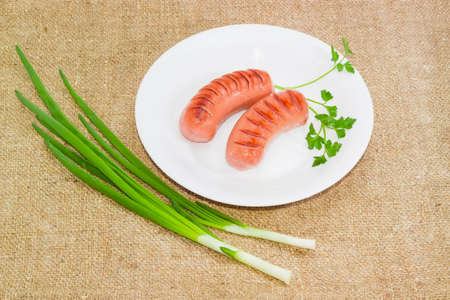 Two grilled thick frankfurters and sprig of parsley on a white dish, green onion on a sackclothの写真素材