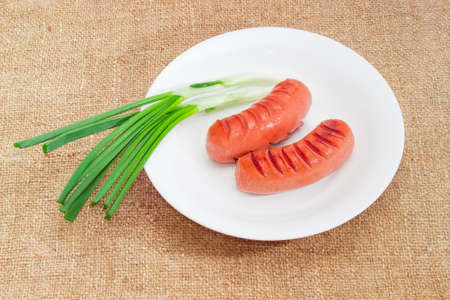 Two fried thick frankfurters and two green onion on a white dish on a sackclothの写真素材