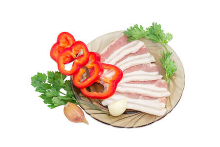 Uncooked slices of streaky pork belly bacon on glass saucer, sliced bell pepper, sprigs parsley and coriander, garlic cloves on a light backgroundの写真素材