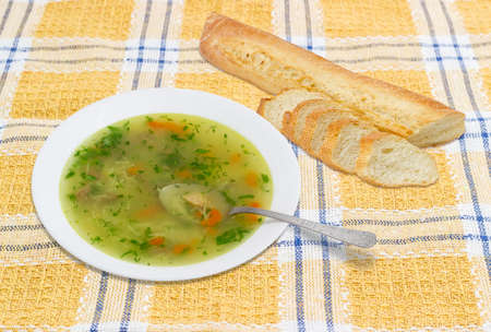 Homemade chicken soup in white dish with a spoon and partly sliced baguette on checkered tableclothの写真素材