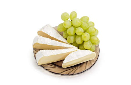 Several slices of Brie cheese and Camembert cheese with cluster of a white table grapes on a dark glass saucer on a light backgroundの写真素材