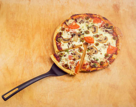 Partly sliced cooked round pizza with chicken, button mushrooms, tomatoes and olives and black plastic spatula on a big wooden cutting boardの写真素材