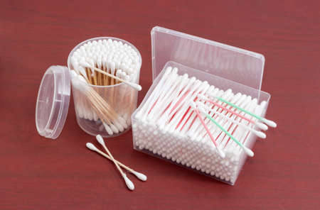 Cotton swabs on a plastic rods in rectangular plastic container, cotton swabs on a wooden rods in round plastic container and several cotton buds separately on a red wooden surface
の写真素材
