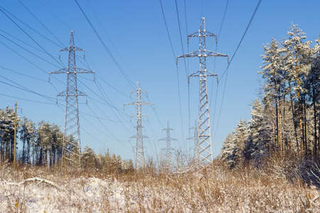 Several overhead power lines with steel lattice structures transmission towers and reinforced concrete poles against the backdrop of sky and pine forest covered snowの写真素材