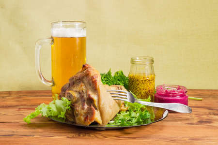 Baked ham hock with lettuce and parsley on a glass dish, glass of lager beer, beet horseradish sauce, French mustard, fork on an old wooden surfaceの写真素材