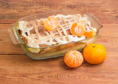 Sweet pancakes filled with cottage cheese with sour cream and sprinkled with cinnamon powder in the rectangular glass roasting pan and mandarins on old wooden surfaceの写真素材