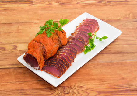 One piece and sliced dried pork tenderloin with twigs of parsley on a white rectangular dish on a wooden surfaceの写真素材