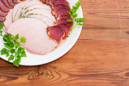 Fragment of white dish with sliced dried pork tenderloin, ham and twigs of parsley closeup on a light background の写真素材