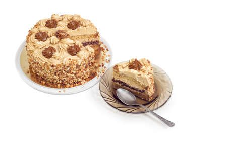 Piece of layered sponge cake, decorated with butter cream, caramelized condensed milk and nuts with spoon on a glass saucer and partly sliced cake on a light backgroundの写真素材
