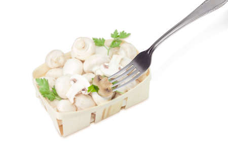 Half of the fried button mushroom with parsley leaf on the stainless steel fork on the background of blurred wooden basket with uncooked mushrooms on a light backgroundの写真素材