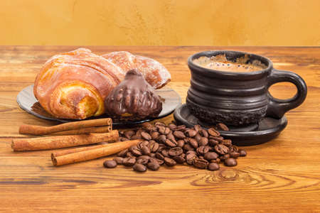 Freshly brewed coffee with milk in the black ceramic cup, scattered beside roasted coffee beans and cinnamon sticks, chocolate truffle and croissant on the glass saucer on the old wooden surface
の写真素材