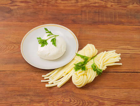 Ball of the fresh mozzarella cheese on the saucer, two portion of the mozzarella cheese in the shape of strings, twisted to form a plait and twigs of parsley on an old wooden surfaceの写真素材