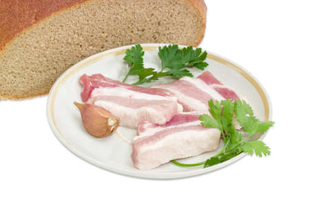Several slices of uncooked streaky pork belly bacon, garlic, twigs of parsley and cilantro on saucer against the background of cut hearth brown bread closeup on a light backgroundの写真素材