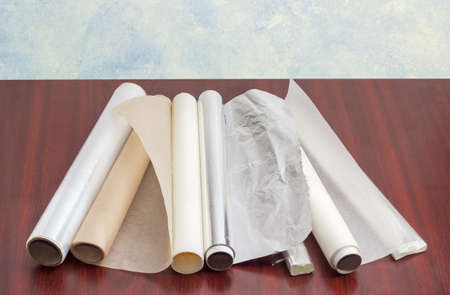 Several rolls of the plastic oven bags, plastic food wrap, aluminum foil and various parchment paper for household use on a dark red wooden surfaceの写真素材