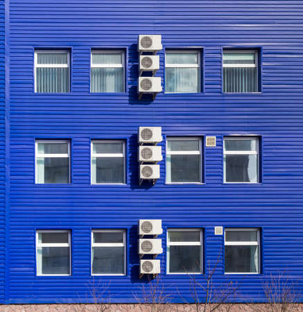 Fragment of the blue wall of a small office building with several window and vertical row of the outside parts of a split-type air conditionersの写真素材
