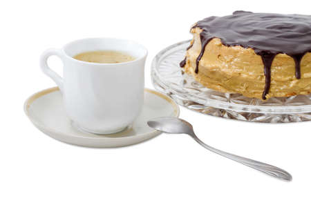 Coffee with cream in the white cup on the saucer with spoon on a background of the sponge cake covered with chocolate glaze closeup on a light backgroundの写真素材